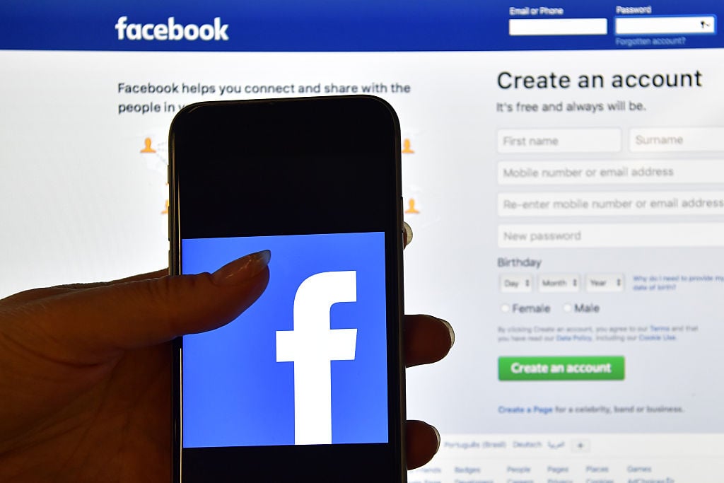 A person holds an iPhone displaying the Facebook app logo in front of a computer screen