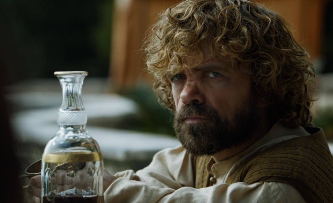 Tyrion scowls with his arms cross, with an empty carafe of wine sitting on the table next to him