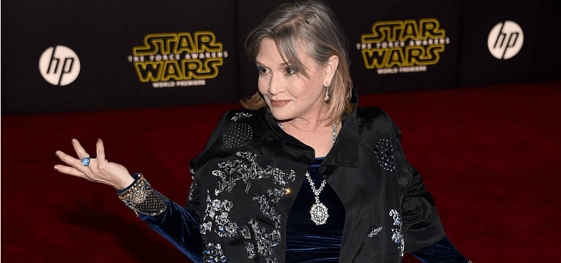 Carrie Fisher has one hand up and is smiling on the Star Wars red carpet.