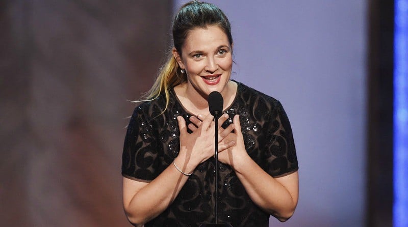 Drew Barrymore onstage in a black dress speaking into a microphone