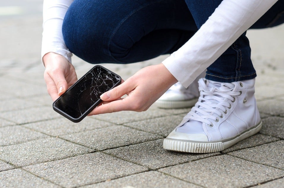 kneeling person in jeans and shoes picking up broken phone