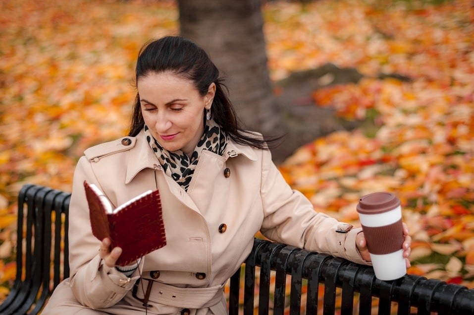 Mature woman sitting on a bench outdoors in a park next to a tree, wearing a trench coat, holding a cup of coffee and reading a book while smiling surrounded by fall foliage in autumn Mature woman sitting on a bench