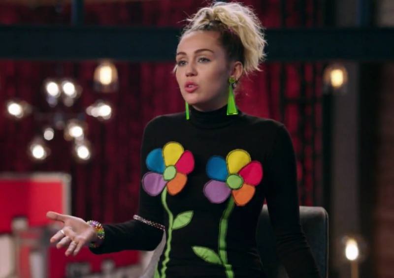 Miley Cyrus is wearing a black turtle neck with two flowers over her chest.