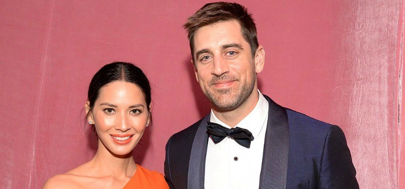 Olivia Munn and Aaron Rodgers standing behind a pink wall.