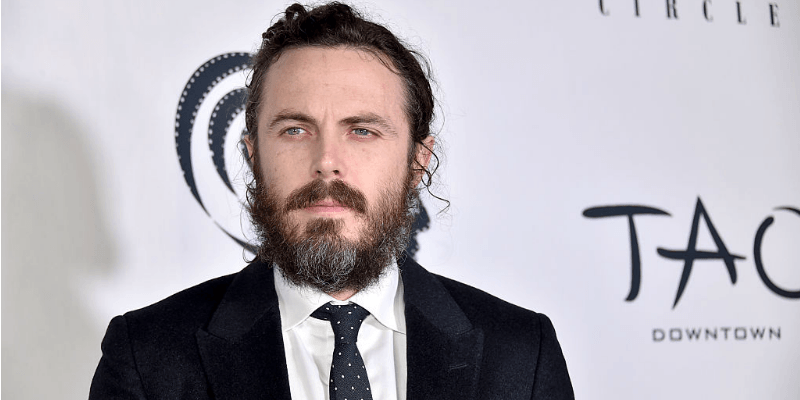Casey Affleck standing on a red carpet in a black suit and tie