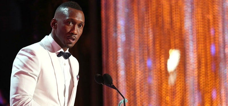 Mahershala Ali speaks into a microphone while on stage in a white tuxedo