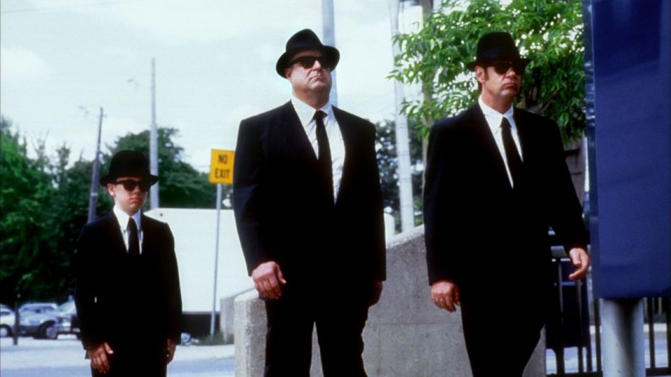 John Goodman and Dan Akroyd wearing black suits, fedoras, and sunglasses
