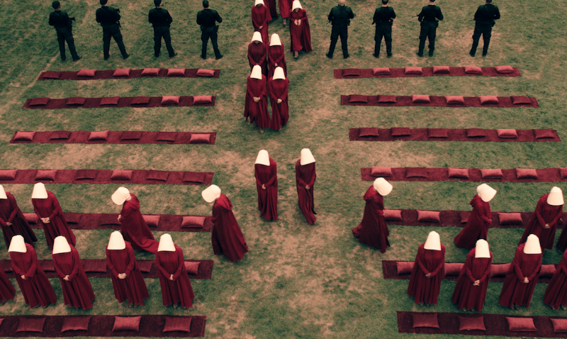 A group of women dressed in red robes with white bonnets known as Handmaids attend an outdoor gathering in Hulu's The Handmaid's Tale