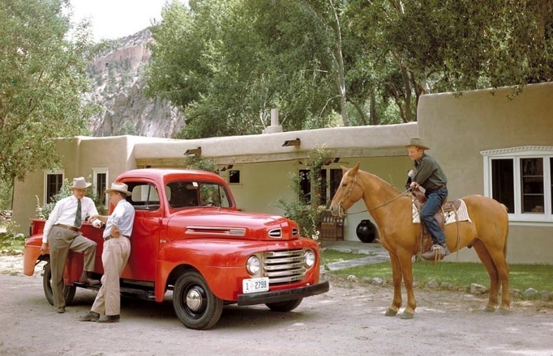 Two men wearing cowboy hats lean on a red Ford F-1 truck as it sits parked in front of a house