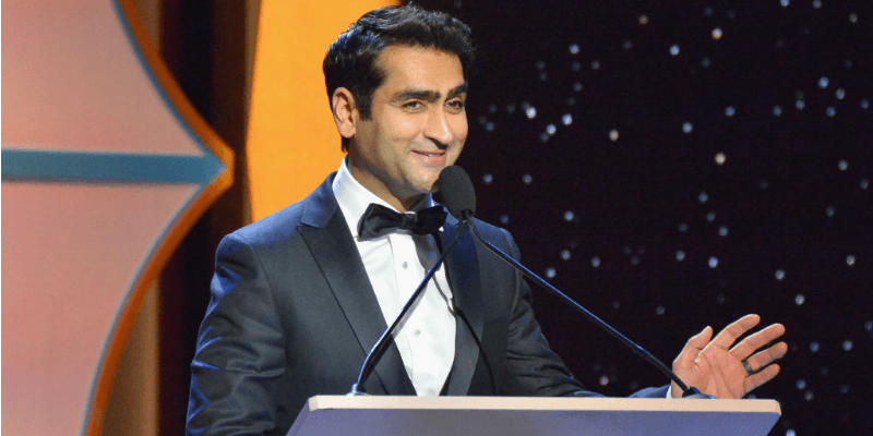 Kumail Nanjiani in a suit and at a podium