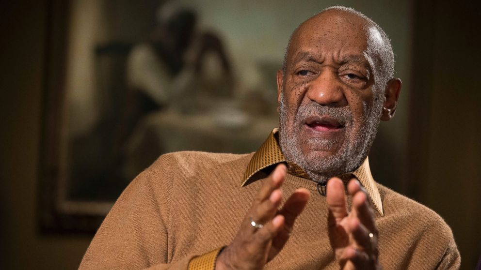 Bill Cosby wearing a tan collared shirt, with his hands out and speaking in an interview