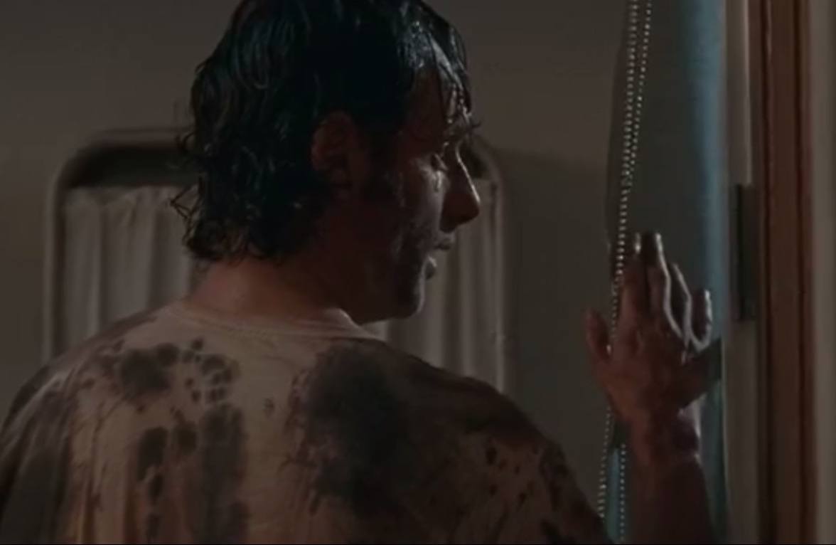 Rick stands in front of a window while wearing a blood-drenched shirt.