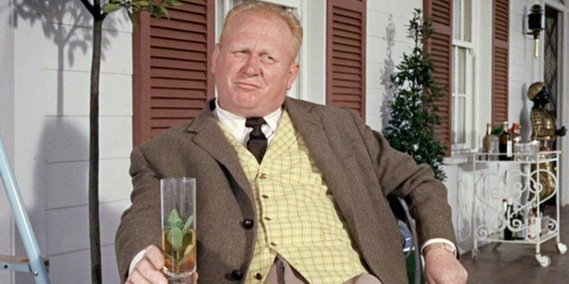 Goldfinger is lounging on a chair on a porch holding a glass.