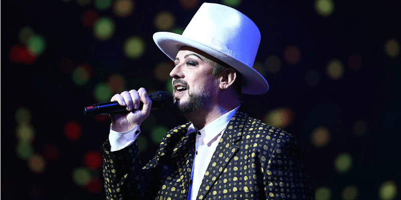 Boy George is wearing a white top hat and in a patterned jacket as he sings.