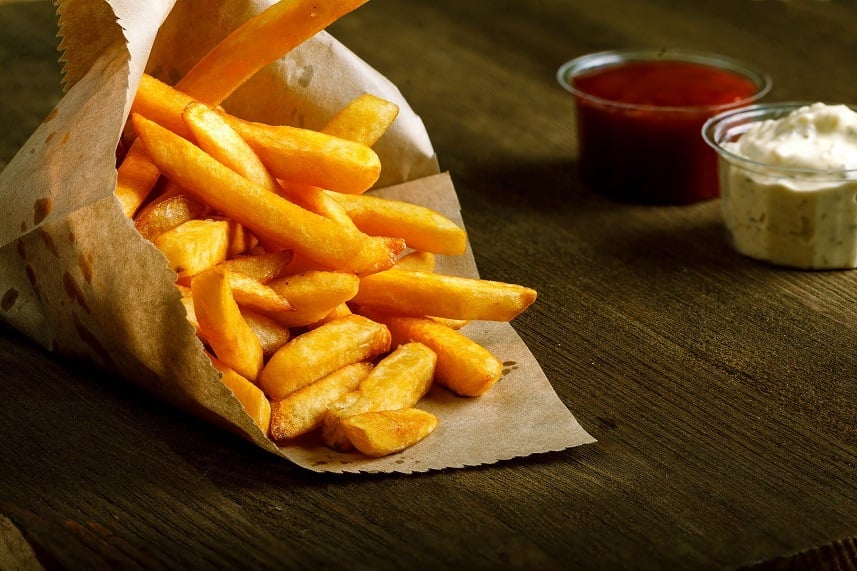 Container of fresh fried french fries with ketchup and white sauce on wooden table.