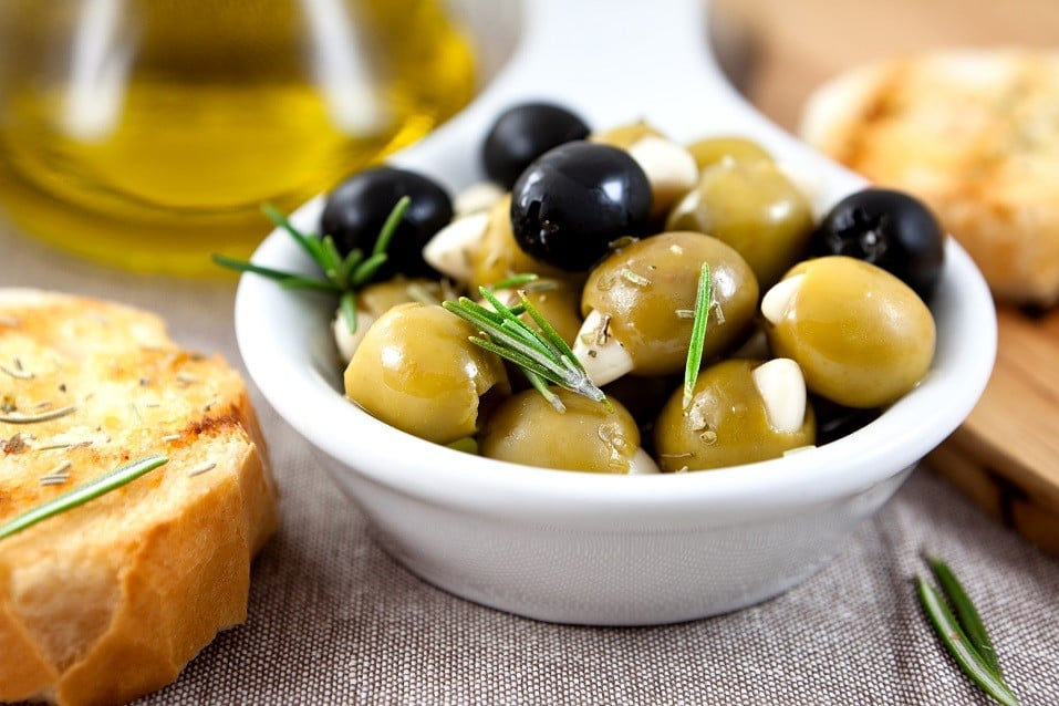 White bowl filled with green and black olives with fresh green herbs on top.