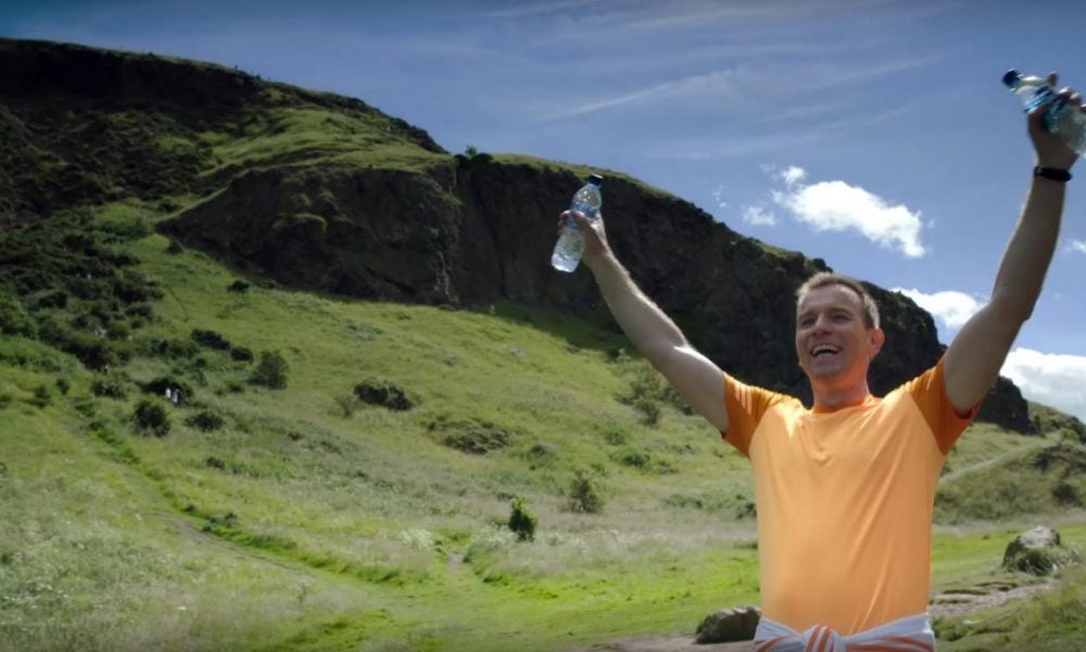 Ewan McGregor with his arms up, and water bottles in either hand, while smiling