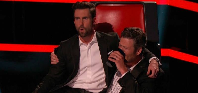Blake Shelton is drinking from a mug while Adam Levine sits in his lap on the set of The Voice