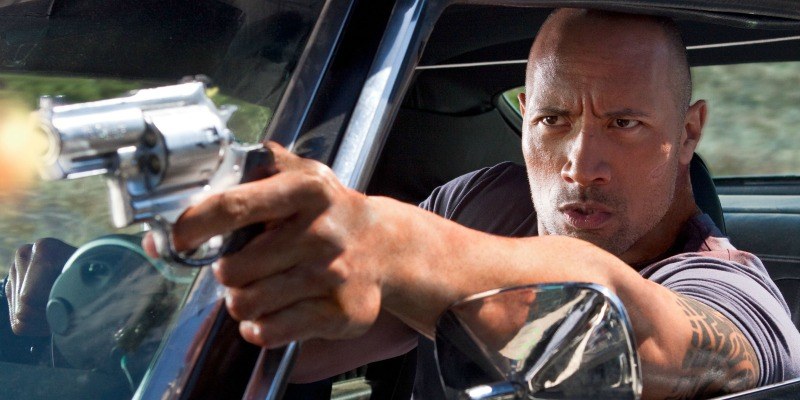 Dwayne Johnson has his arm out of a car pointing a gun.