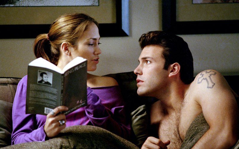 Jennifer Lopez reads a book in bed, while a shirtless Ben Affleck looks up at her from a prone position
