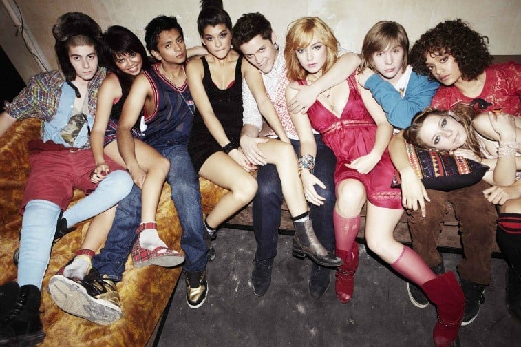 Nine teenagers with their arms around each other on a couch in a dingy room