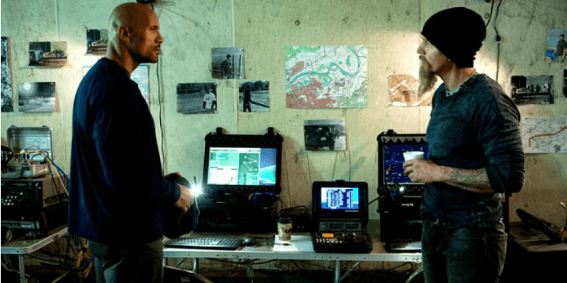 Dwayne Johnson and another man are talking in a room of computers in Snitch.