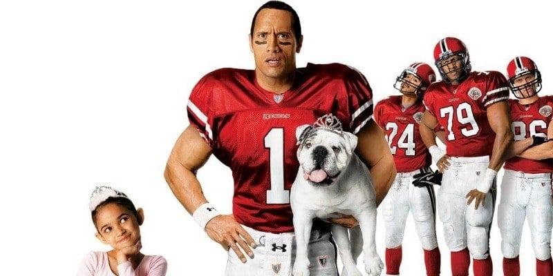 Dwayne Johnson poses in a football uniform holding a dog. A little girl is smiling on his left and his football teammates are on his right.