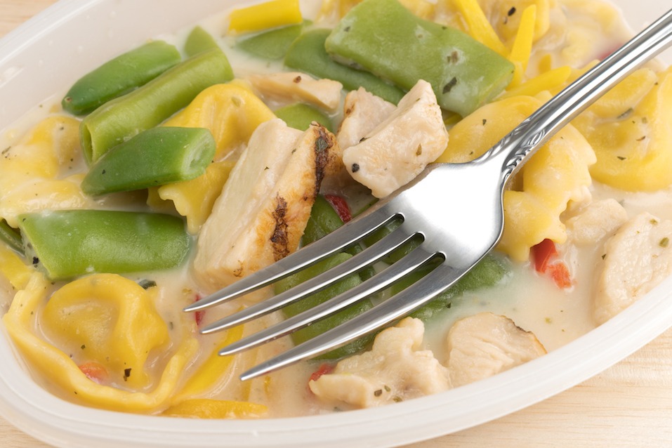 Chicken with tortellini and vegetable TV dinner in a plastic tray with a fork atop a wood table.
