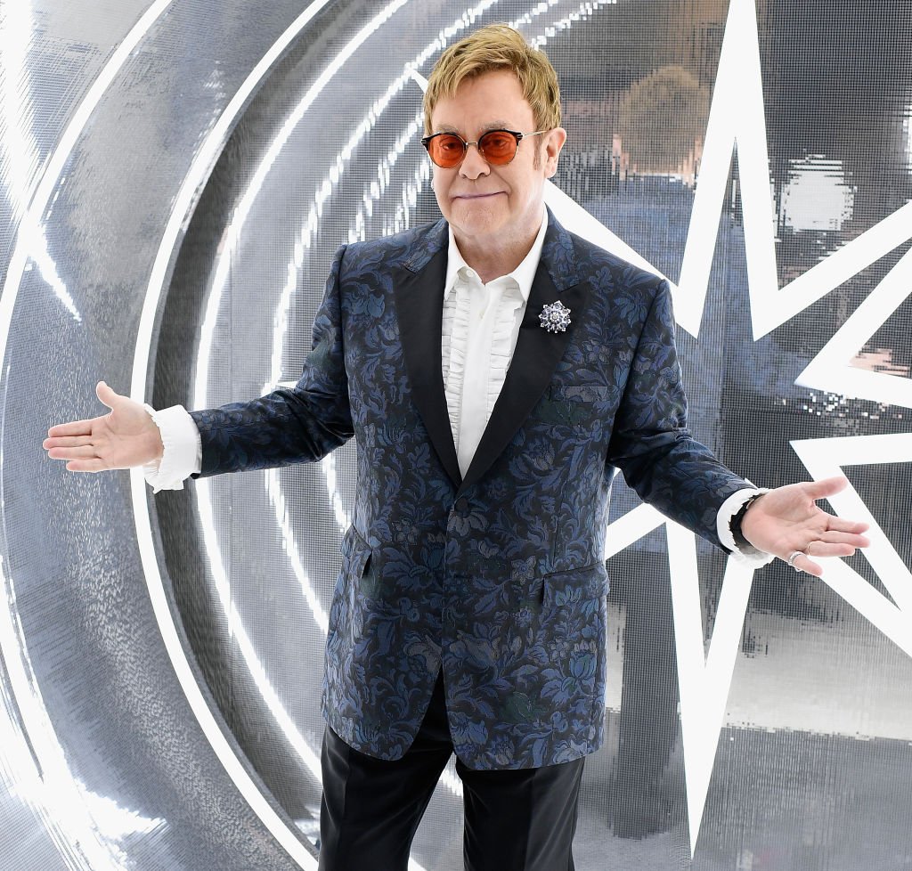 Elton John posing in front of a bright silver wall.