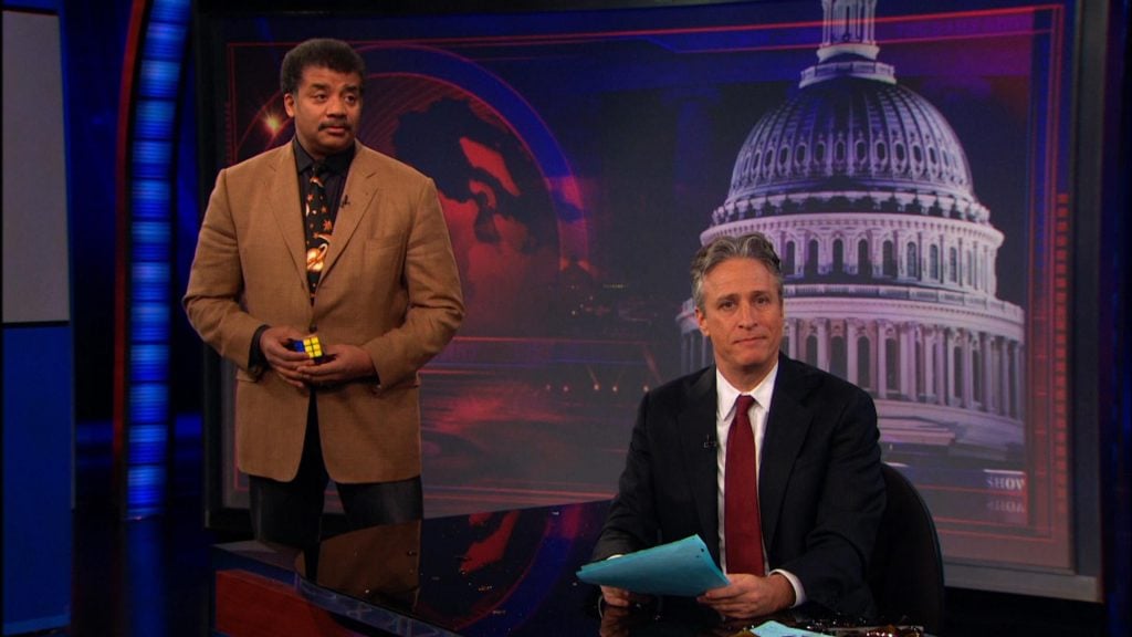 Neil Degrasse Tyson holding a Rubiks Cube, standing behind Jon Stewart on The Daily Show
