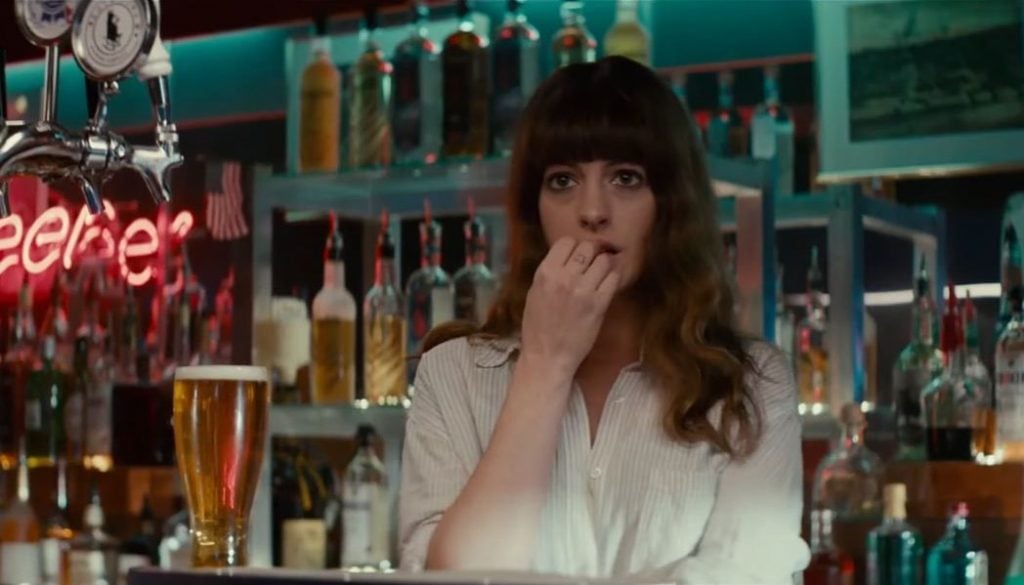 Anne Hathaway behind a bar next a beer, with her hand up to her mouth