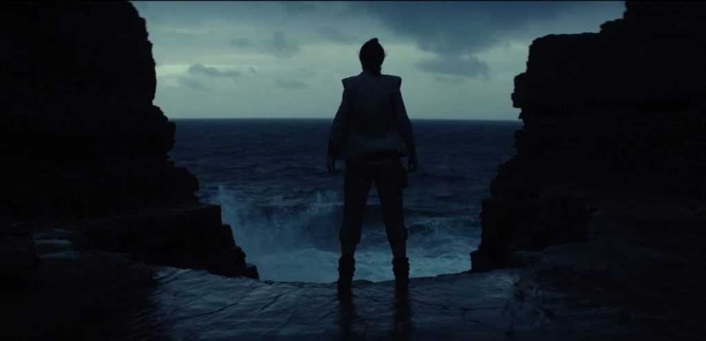Rey stands in front of a cliff face, with waves crashing in front of her