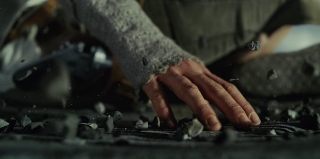 Rey with one hand placed on the ground, lifting a group of pebbles together with the Force