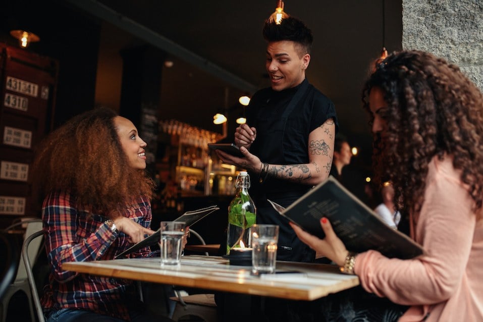 Two women sitting at cafe holding menu card giving an order to male waiter.