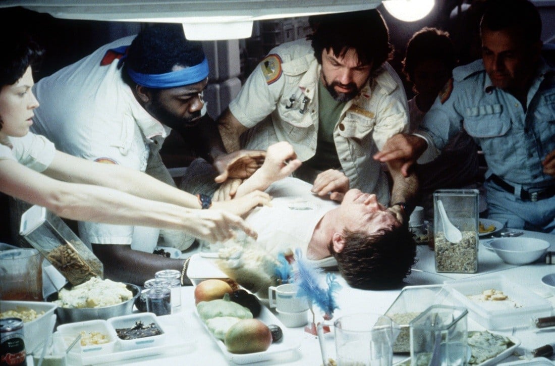 Actors gather around John Hurt, who's lying on a table wearing a white tshirt