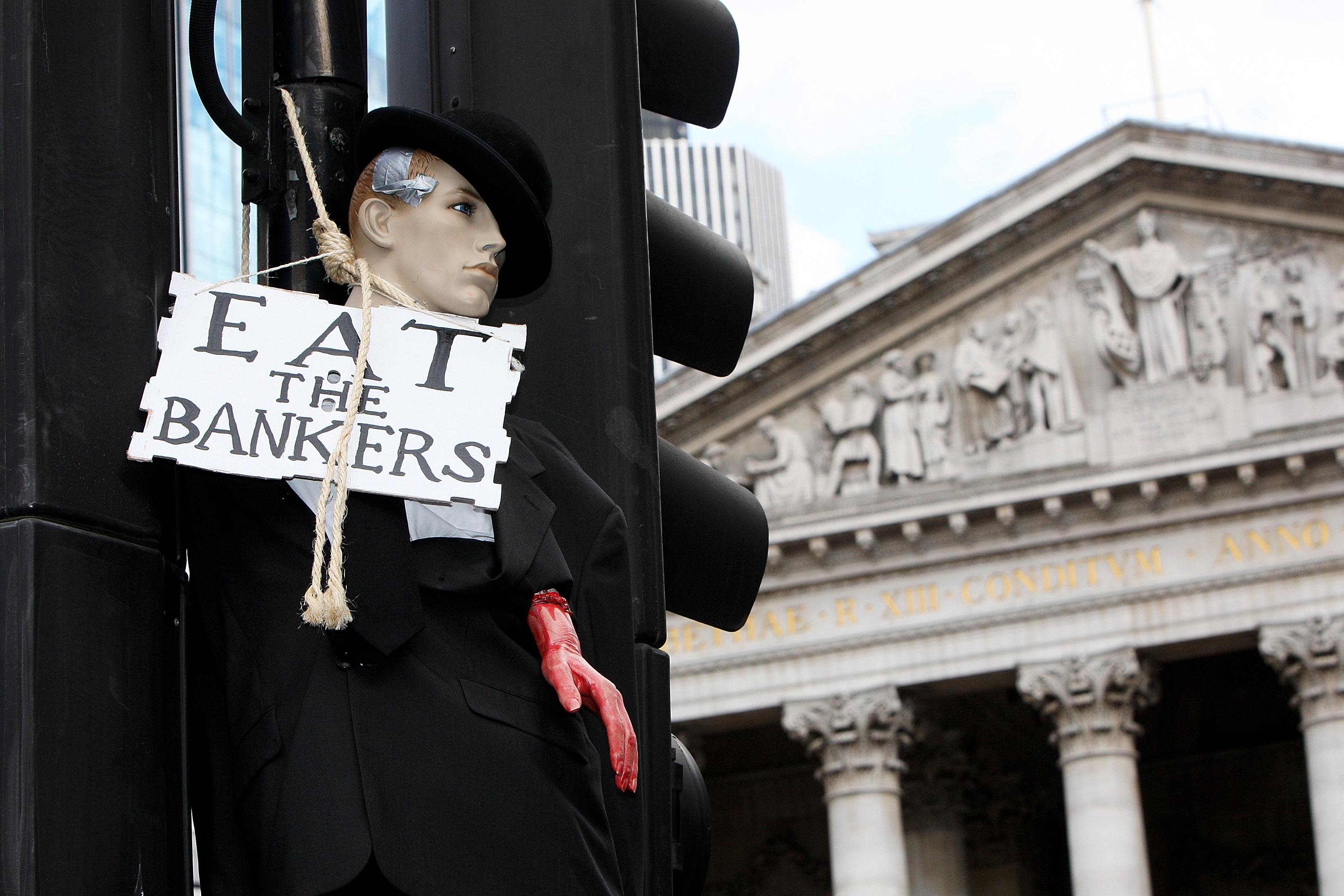 A mannequin dressed as a banker hangs from a traffic light post with a sign that says "eat the bankers."