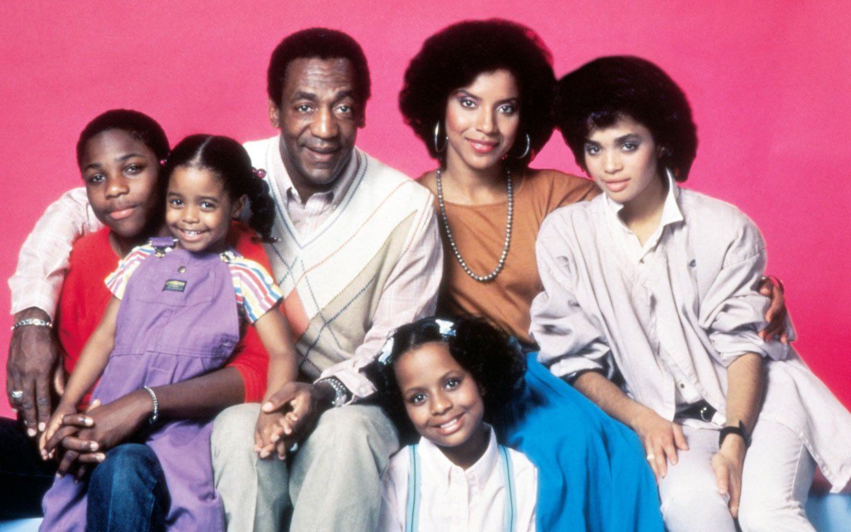 The cast of the The Cosby Show poses together in front of a pink background