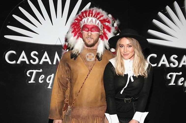 Trainer Jason Walsh in a Native American costume standing with girlfriend Hilary Duff in a pilgrim costume in front of a back wall that reads 'Casamigos Tequila'