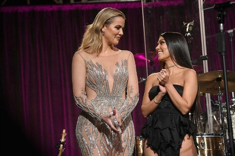 Khloe and Kourtney Kardashian onstage in fancy attire in front of musical instruments and a fuchsia curtain