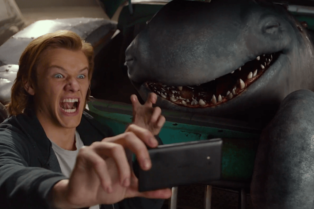Lucas Till with his mouth open, taking a selfie with a shark-like monster