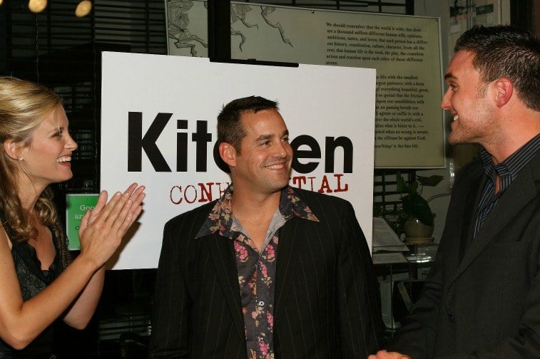 Two men and a woman speaking to one another in front of a sign for 'Kitchen Confidential'