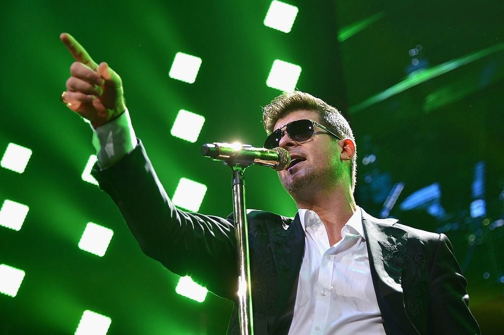 Singer Robin Thicke on stage wearing sunglasses and singing into a microphone while pointing