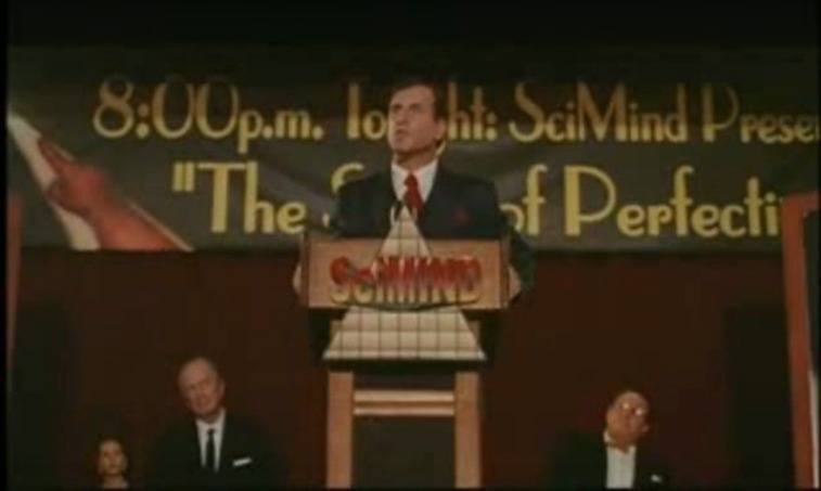 Man in a suit standing at a podium with a banner and other men behind him in The Profit