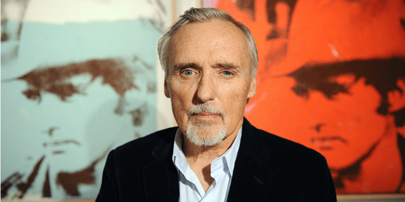 Dennis Hopper poses in front of two photos of him in a cowboy hat.