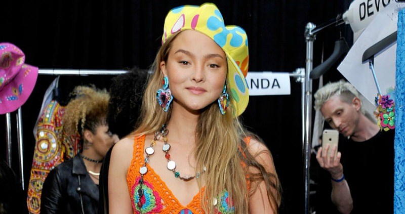 Devon Aoki is in a bright colored dress and hat backstage at a fashion show.