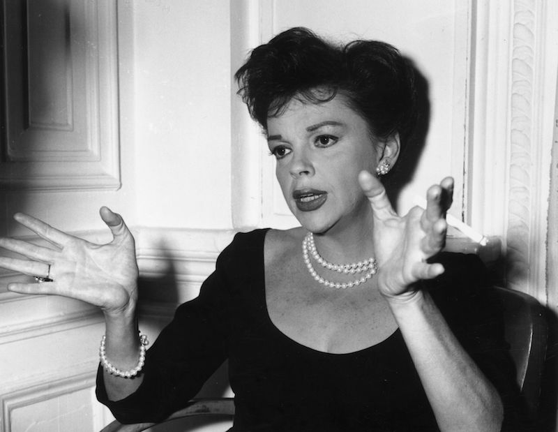 Judy Garland gesture with her hands while wearing a black dress and pearls