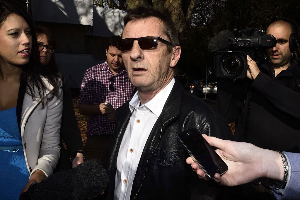 Phil Rudd wearing sunglasses and a suit, speaking into a collection of microphones