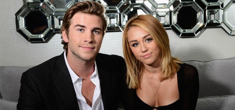Miley Cyrus and Liam Hemsworth are sitting together and smiling.