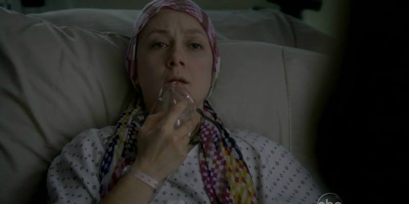 Sara Gilbert has a head scarf on her head and is in a hospital gown. She is pulling an oxygen mask down from her mouth.