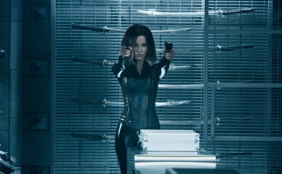 Kate Beckinsale in a black leather suit, holding our twin guns, standing against a rack of swords